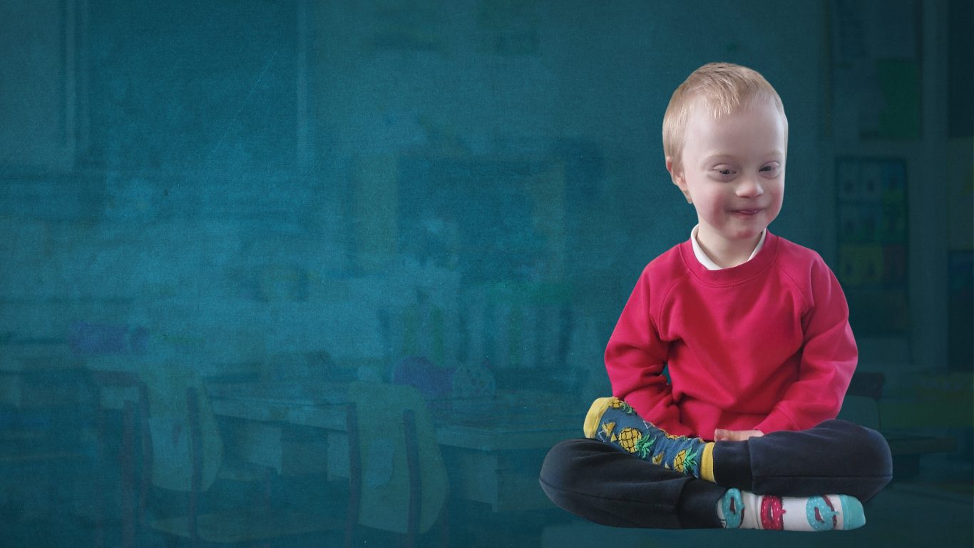 Young child with down syndrome in primary school uniform sitting cross legged and smiling. He's not wearing shoes and his socks are unmatched and whacky colours