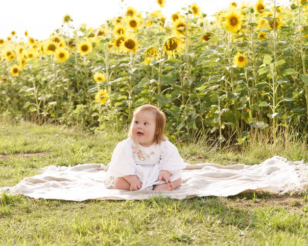 Baby in a white gown laying on a white blanket on a sunny day. She's on the grass and behind her is a field of giant sunflowers!