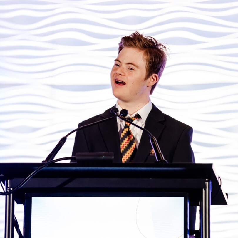 DSUK Patron Noah Matthews Matofsky standing on stage and delivering a talk in a suit at a conference