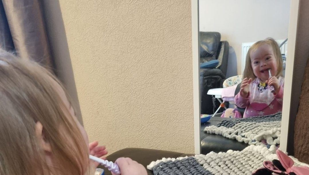 Toddler with down syndrome is cleaning her teeth in the mirror and smiling as if shes really enjoying the experience