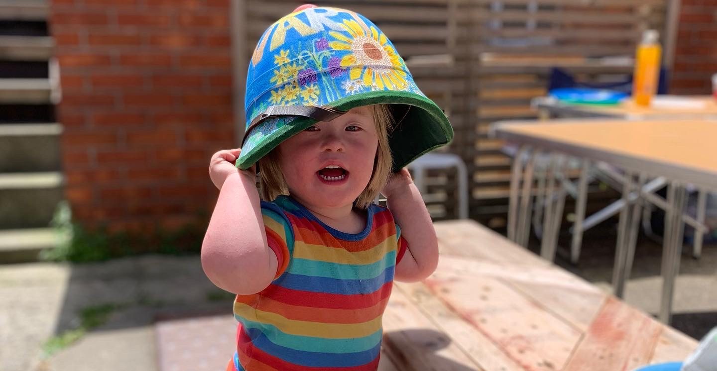 Child with down syndrome is putting on an adult sized hat while staring at the camera and posing for a photo