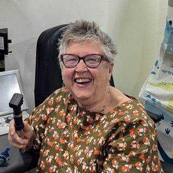 Maggie woodhouse is sitting in front of a busy desk and computer, facing sideways and looking at a camera smiling