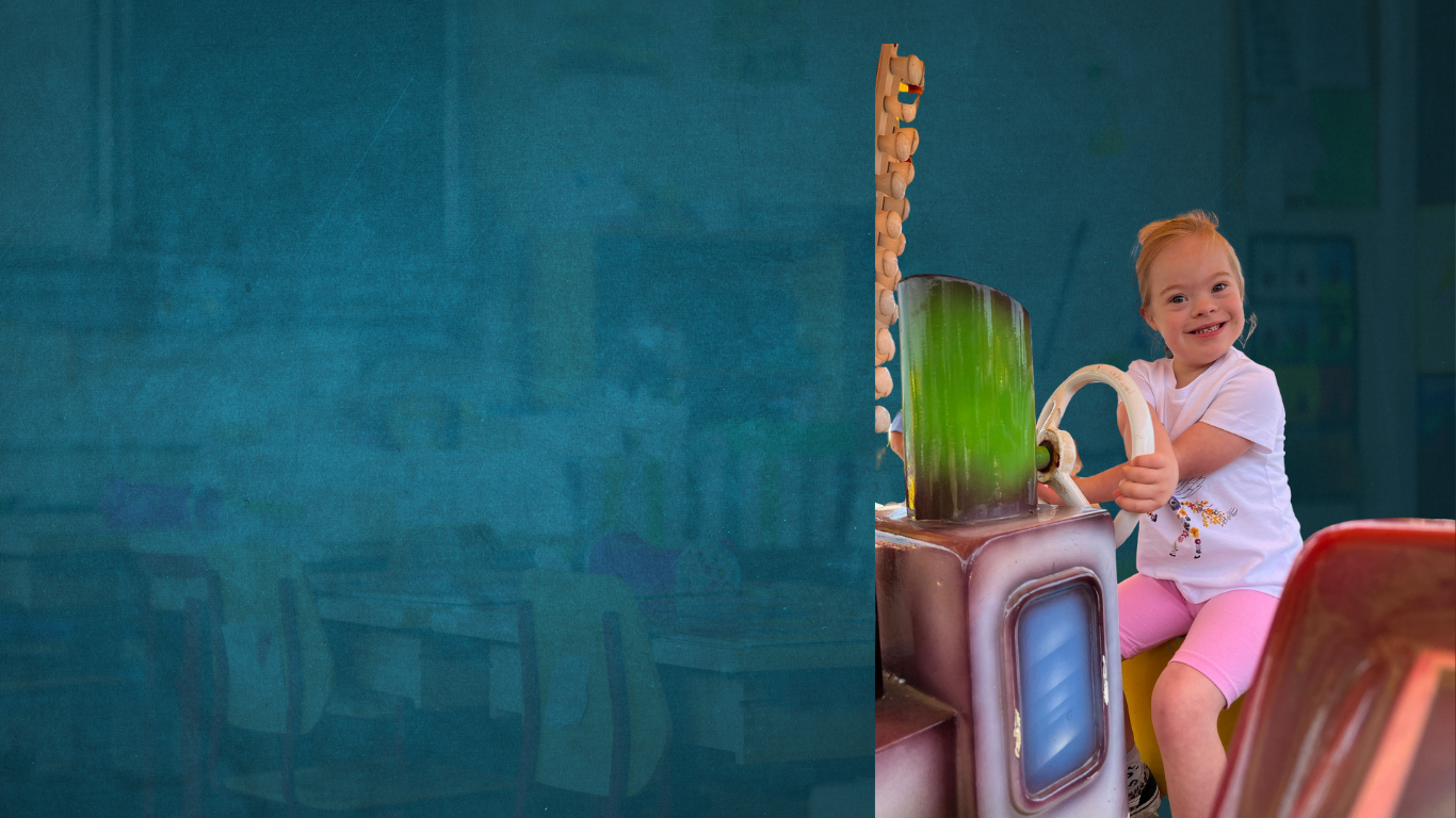 Young child with down syndrome is on a merry go round toy car holding the wheel and smiling for the camera